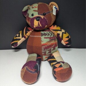 May include: A plush teddy bear constructed from various fabric patches featuring geometric patterns and solid colors including brown, sage green, orange, and purple. The bear has black button eyes, a stitched nose, and a small white tag on its ear.