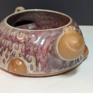 May include: A handmade ceramic bowl with a unique design. The bowl features a reddish-purple glaze with tan accents and a textured pattern. It has two brown, rounded handles and a small, circular opening. The bowl is likely used for decorative purposes.