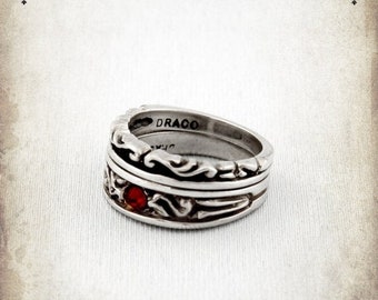 May include: Silver ring with intricate scrollwork and a red gemstone. The ring is engraved with the word "DRACO".