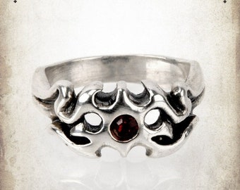 May include: Silver ring with a red gemstone set in a detailed, intricate design. The ring is a statement piece with a gothic style.