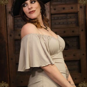 May include: A beige off-the-shoulder top with flared sleeves. The image also features a black pirate hat with lace trim. The background is a dark wooden door. A small emblem is visible at the bottom.