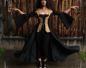 Puede incluir: Una mujer con un vestido gótico negro y dorado con mangas largas y un corpiño tipo corsé. El vestido se combina con pantalones y botas negras. El fondo es una puerta de madera. La palabra "Bracolite" está en la parte superior.