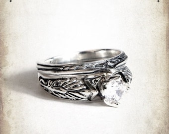 May include: A silver ring with a clear gemstone and intricate leaf-like designs. The ring is set against a white background.