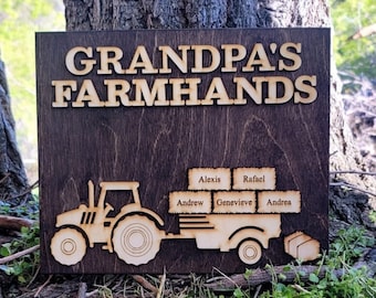 Könnte beinhalten: Ein Holzschild mit dem Text "Grandpa's Farmhands" und einer Silhouette eines Traktors, der einen Anhänger zieht. Die Namen Alexis, Rafael, Andrew, Genevieve und Andrea sind auf dem Anhänger geschrieben.