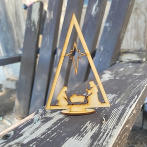 May include: A wooden Christmas nativity scene. The scene is cut out of light-colored wood and features a star, Mary, Joseph, and baby Jesus. The figures are inside a triangle frame and sit on a round base. The background is a weathered wooden chair.