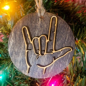 May include: A wooden Christmas ornament with a dark brown stain. The ornament features a cutout design of the American Sign Language symbol for "I love you".