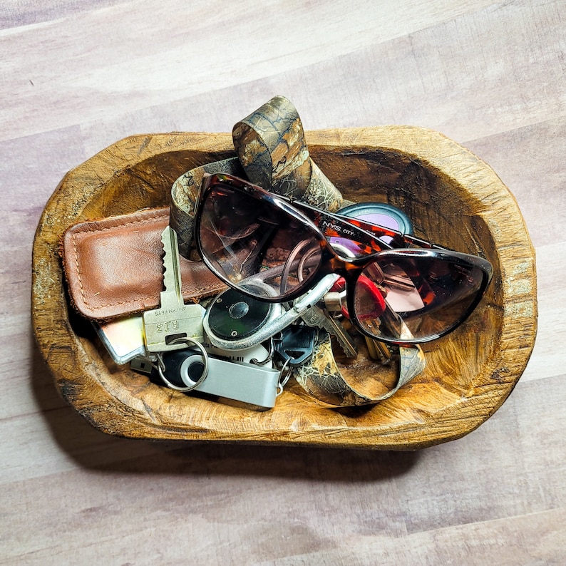 May include: A wooden bowl filled with various items, including a pair of brown sunglasses, a leather wallet, keys, and a small metal tool. The bowl is on a light wood surface.
