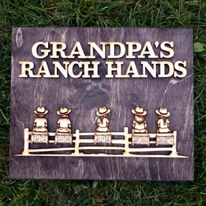 May include: Wooden sign with the words "GRANDPA'S RANCH HANDS" in raised letters. Below, five figures in cowboy hats sit on a fence, each with a name tag: Dakoda, Daxdan, Allysa, Abrianna, and Annabeth.