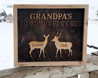 May include: A wooden sign with the text "Grandpa's Hunting Buddies" and two deer silhouettes, one labeled "Eleonore" and the other "Logan".