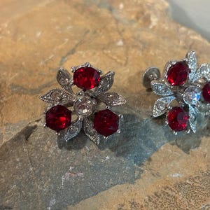 May include: A pair of silver-toned metal screw-back earrings featuring a floral design. Each earring is set with three round, deep red faceted stones and smaller clear accent stones arranged among textured metal leaves and petals.