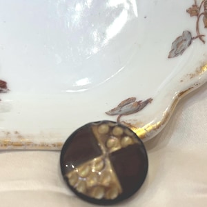 Puede incluir: Un botón redondo de color marrón oscuro con un diseño geométrico dorado y blanco. El botón tiene una superficie texturizada en relieve.