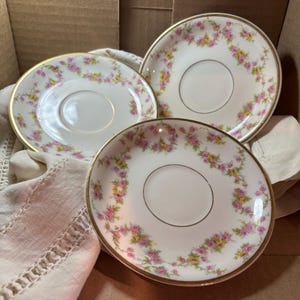 May include: Three white porcelain saucers with a gold rim and a floral pattern of pink and yellow roses. The saucers are arranged on a white linen cloth inside a cardboard box. The saucers are approximately 5 inches in diameter.