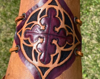 May include: A brown leather archery bracer with a decorative cross design in black and purple. The bracer is laced with leather cord and is held in front of a wooden bow.