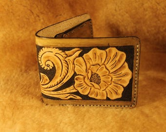 May include: A bi-fold leather wallet with a floral design. The wallet features a tan flower and scrollwork on a black background. The edges are tan with visible stitching. The wallet is open, revealing the interior.