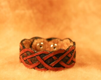 May include: A handcrafted leather bracelet with a woven design. The bracelet features a dark brown base with red interwoven straps. Two silver-colored snaps are visible on the inside of the bracelet. The background is a solid orange color.