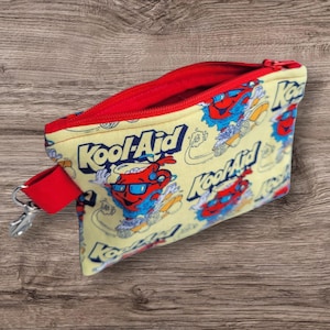 Puede incluir: Una bolsa amarilla con cremallera, interior rojo y correa de mu&ntilde;eca roja. La bolsa tiene un estampado del personaje Kool-Aid Man y el logotipo de Kool-Aid en rojo, azul y blanco. Un cierre plateado est&aacute; unido a la correa de mu&ntilde;eca.
