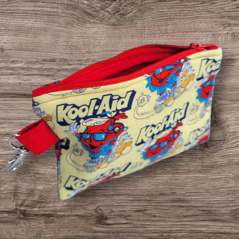 Puede incluir: Una bolsa amarilla con cremallera, interior rojo y correa de mu&ntilde;eca roja. La bolsa tiene un estampado del personaje Kool-Aid Man y el logotipo de Kool-Aid en rojo, azul y blanco. Un cierre plateado est&aacute; unido a la correa de mu&ntilde;eca.