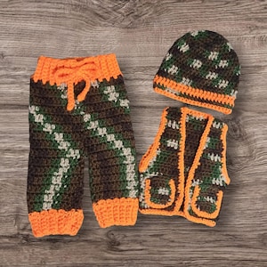 May include: A three-piece crochet baby outfit in a camouflage pattern. The set includes trousers with an orange drawstring, a waistcoat, and a beanie. The trim on all three pieces is bright orange.