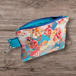 Puede incluir: Peque&ntilde;o estuche abierto con estampado floral, cremallera turquesa y correa de mu&ntilde;eca turquesa con cierre de plata. El estuche presenta un colorido estampado floral con flores rosas, naranjas y amarillas sobre fondo azul.