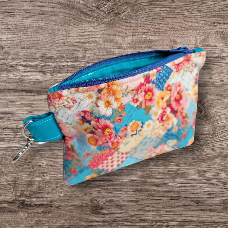 Puede incluir: Peque&ntilde;o estuche abierto con estampado floral, cremallera turquesa y correa de mu&ntilde;eca turquesa con cierre de plata. El estuche presenta un colorido estampado floral con flores rosas, naranjas y amarillas sobre fondo azul.