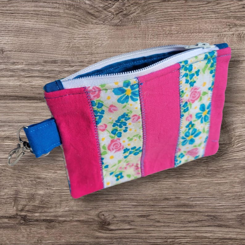 Puede incluir: Una bolsa con cremallera hecha a mano con un dise&ntilde;o vibrante. La bolsa presenta rayas verticales alternas de tela rosa intenso y tela estampada floral con flores azules y rosas sobre un fondo blanco. Se adjunta una correa de mu&ntilde;eca azul y un cierre plateado.