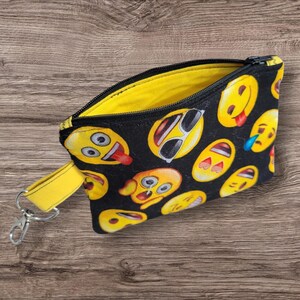Puede incluir: Peque&ntilde;o estuche negro con cremallera, interior amarillo y correa de mu&ntilde;eca amarilla. El exterior est&aacute; cubierto de caras emoji amarillas sobre un fondo negro. Los emojis tienen varias expresiones, incluyendo gafas de sol, l&aacute;grimas y lenguas.