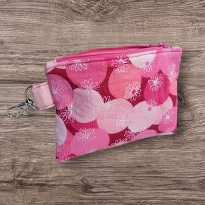 Puede incluir: Un peque&ntilde;o monedero rosa con estampado floral con cremallera a juego y cierre plateado. El bolso presenta un patr&oacute;n repetido de contornos de flores blancas sobre un fondo de c&iacute;rculos rosas y rojos.