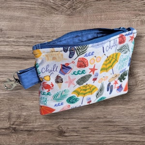 Puede incluir: Peque&ntilde;a bolsa con cremallera abierta, interior azul y correa de mu&ntilde;eca azul con cierre plateado. El exterior blanco est&aacute; estampado con ilustraciones de tem&aacute;tica playera, incluyendo sombrillas, gafas de sol y conchas marinas.