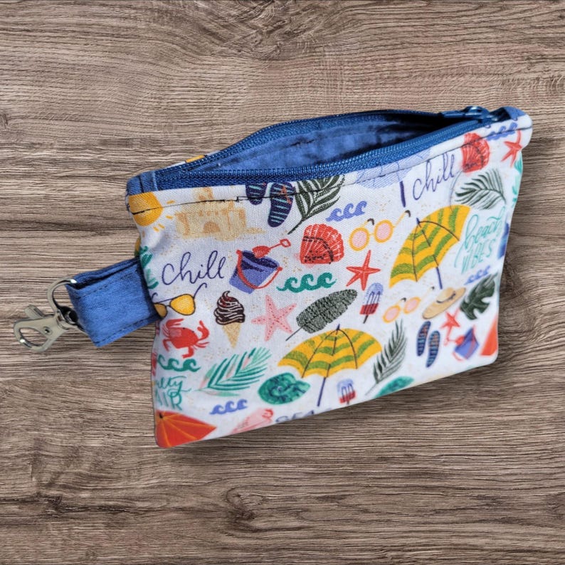 Puede incluir: Peque&ntilde;a bolsa con cremallera abierta, interior azul y correa de mu&ntilde;eca azul con cierre plateado. El exterior blanco est&aacute; estampado con ilustraciones de tem&aacute;tica playera, incluyendo sombrillas, gafas de sol y conchas marinas.