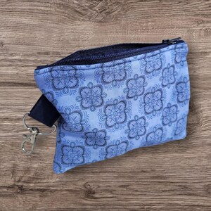 Puede incluir: Una peque&ntilde;a bolsa con cremallera azul claro con un estampado floral repetido en azul m&aacute;s oscuro. La bolsa tiene una cremallera azul marino y una correa azul marino con un cierre plateado. La bolsa est&aacute; sobre una superficie de madera.