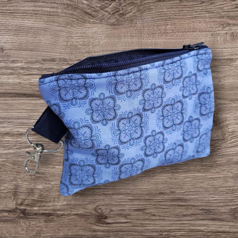 Puede incluir: Una peque&ntilde;a bolsa con cremallera azul claro con un estampado floral repetido en azul m&aacute;s oscuro. La bolsa tiene una cremallera azul marino y una correa azul marino con un cierre plateado. La bolsa est&aacute; sobre una superficie de madera.