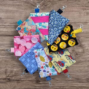 Puede incluir: Una colecci&oacute;n de peque&ntilde;as bolsas de tela coloridas con varios dise&ntilde;os, incluyendo florales, emoji y geom&eacute;tricos. Cada bolsa tiene un clip met&aacute;lico y un lazo de tela. Las bolsas est&aacute;n dispuestas en un patr&oacute;n circular sobre una superficie de madera.