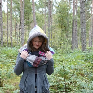 May include: A woman wearing a grey hooded jacket with a colourful patchwork scarf in a woodland setting.