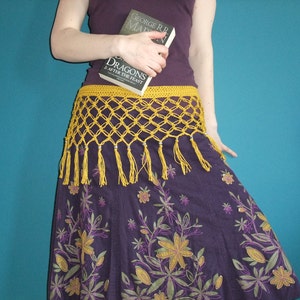 May include: A woman wearing a purple skirt with floral embroidery and a yellow crocheted belt with tassels. She is holding a book titled "Dragons: After the Feast" by George R.R. Martin.