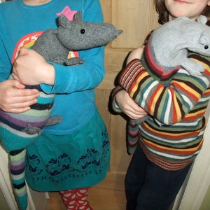 May include: Two children are holding grey and white striped fabric animal toys. The child on the left is wearing a blue shirt and a teal skirt. The child on the right is wearing a striped jumper and blue trousers.