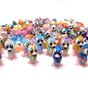 May include: A collection of colorful, small bird figurines. The birds have large, round eyes and pink feet. The figurines are in various colors, including blue, yellow, orange, and purple. The birds are standing on a white surface.