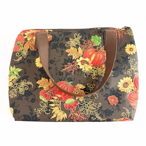 May include: A rectangular tote bag with a brown background and a fall-themed pattern. The pattern includes orange pumpkins, yellow sunflowers, and red berries. The bag has brown handles and a zipper closure.