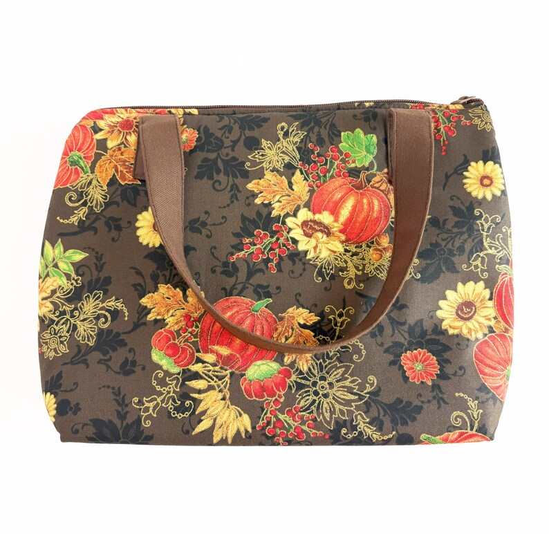 May include: A rectangular tote bag with a brown background and a fall-themed pattern. The pattern includes orange pumpkins, yellow sunflowers, and red berries. The bag has brown handles and a zipper closure.