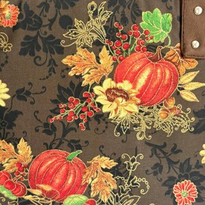 May include: Close-up of a fabric with a fall harvest theme. The design features orange pumpkins, sunflowers, red berries, and autumn leaves. The background is a dark brown with a black floral pattern, and gold accents.