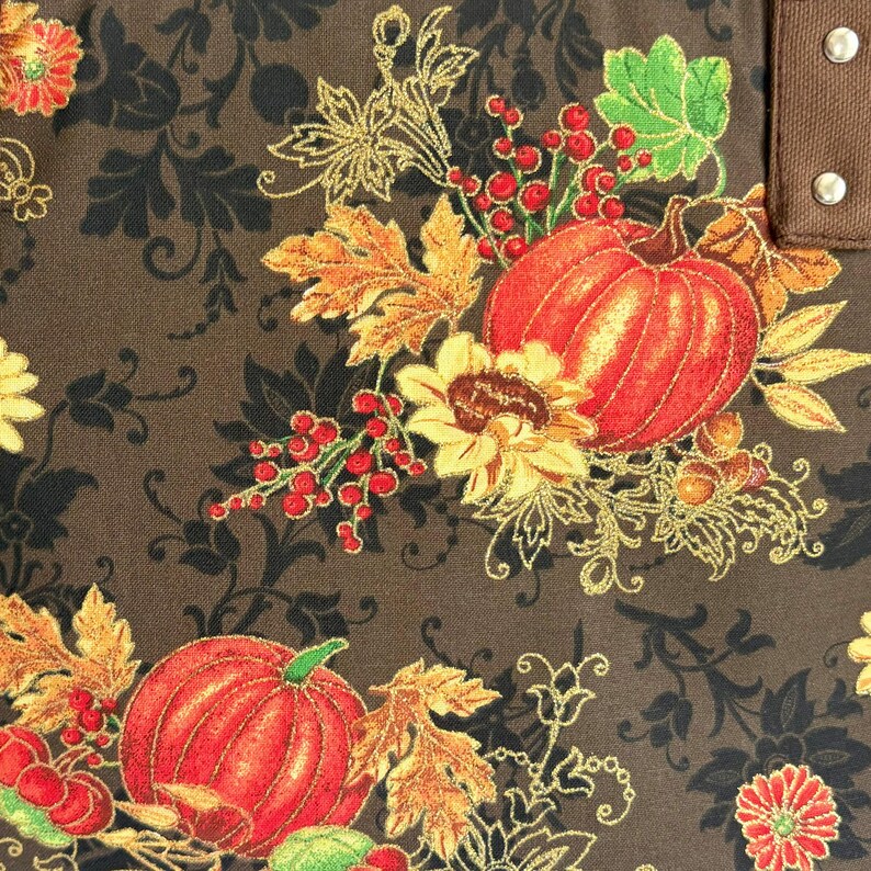 May include: Close-up of a fabric with a fall harvest theme. The design features orange pumpkins, sunflowers, red berries, and autumn leaves. The background is a dark brown with a black floral pattern, and gold accents.