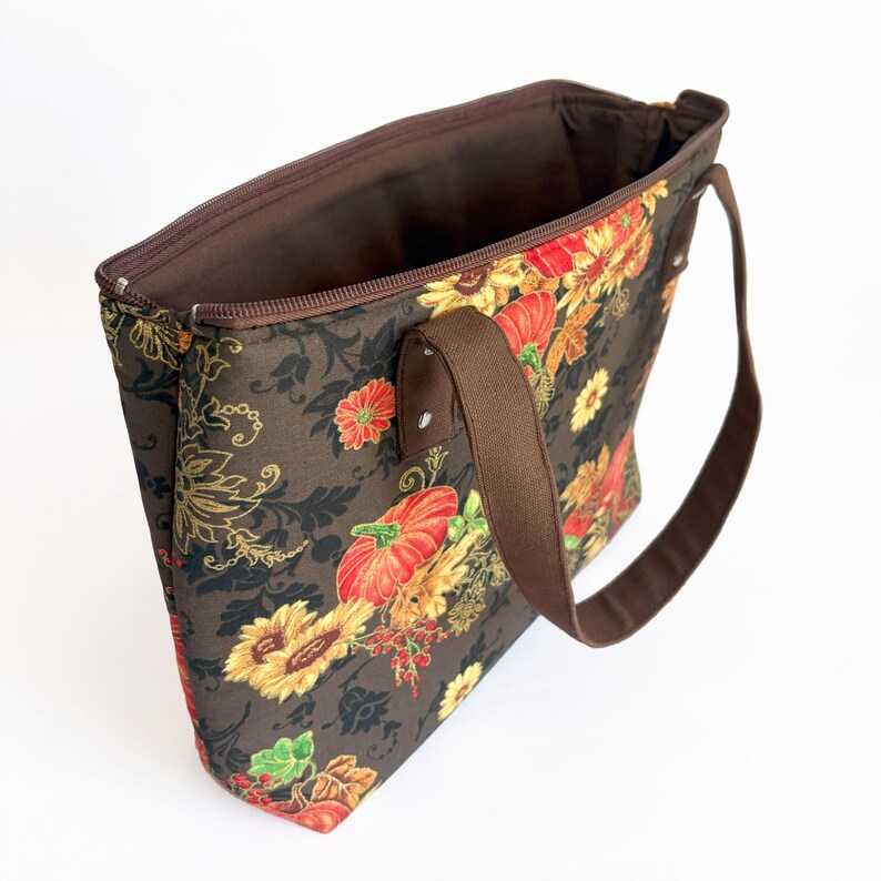 May include: A brown tote bag with a floral pattern featuring sunflowers, pumpkins, and other autumn elements. The bag has a brown zipper, handles, and interior lining. The bag is open, showing the interior.