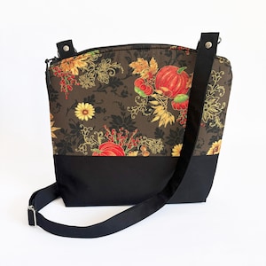 May include: A crossbody bag with a black base and strap. The upper portion features a brown floral pattern with pumpkins, sunflowers, and fall foliage. The bag has a zipper closure and adjustable strap.