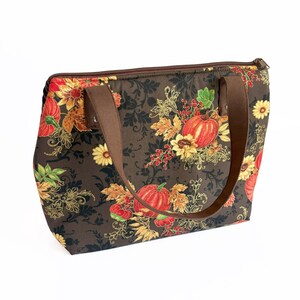 May include: A brown tote bag with a fall-themed floral pattern featuring pumpkins, sunflowers, and leaves in shades of red, orange, and yellow. The bag has brown handles and a zipper closure. The bag is designed for carrying items.