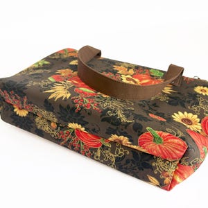 May include: A brown tote bag with a fall-themed floral pattern featuring pumpkins, sunflowers, and foliage in shades of red, orange, yellow, and green. The bag has a brown handle and a rectangular shape.