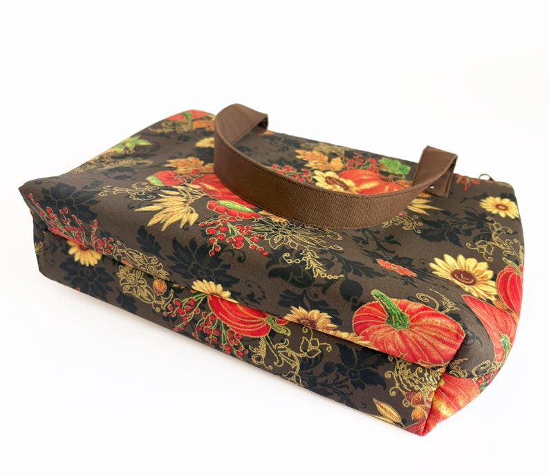 May include: A brown tote bag with a fall-themed floral pattern featuring pumpkins, sunflowers, and foliage in shades of red, orange, yellow, and green. The bag has a brown handle and a rectangular shape.