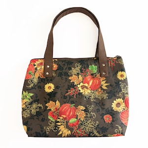 May include: A brown tote bag with a fall-themed floral pattern featuring pumpkins, sunflowers, and autumn leaves. The bag has brown handles and a zipper closure. The bag is designed for carrying items.