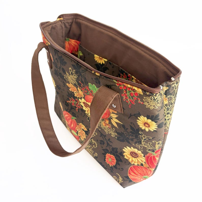 May include: Brown tote bag with a floral pattern featuring pumpkins, sunflowers, and fall foliage. The bag has a brown handle and a zipper closure. The interior is brown with pockets.
