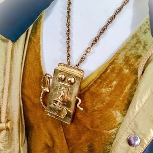 May include: A gold chain necklace with a vintage-style phone charm. The phone charm has a rotary dial and a receiver.