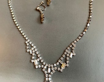 Op de afbeelding: Een zilverkleurige ketting en bijpassende oorbellen set. De ketting heeft een reeks kleine, heldere strass-steentjes en een centraal hangerontwerp. De oorbellen zijn in schroefstijl, met een gebogen ontwerp van strass-steentjes.