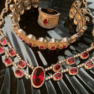 Puede incluir: Un conjunto de tres piezas de joyería de tono dorado con piedras preciosas rojas. El conjunto incluye un collar, una pulsera y un anillo. El collar y la pulsera presentan una doble fila de piedras preciosas rojas de forma ovalada con diamantes más pequeños que las rodean. El anillo tiene una sola piedra preciosa roja de forma ovalada con diamantes que la rodean.